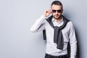 In his own perfect style. Handsome young man in smart casual wear adjusting his sunglasses and looking at camera while standing against grey background photo