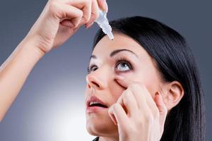 Taking care of her vision. Side view of young women applying eye drops while standing against grey background photo