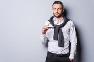 Charming handsome. Handsome young man in in smart casual wear holding sunglasses and looking at camera while standing against grey background photo