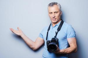 aquí puede ser su publicidad. retrato de un hombre maduro confiado en una camiseta sosteniendo la cámara y señalando mientras está de pie contra un fondo gris foto