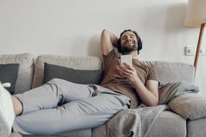 Cheerful young man in headphones holding smart phone while relaxing on the couch at home photo