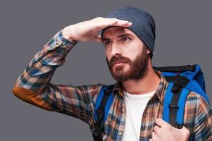 Exploring new places. Handsome young bearded man carrying backpack and looking at view while holding hand upon his eyes and standing against grey background photo