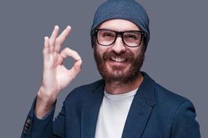 Everything is OK. Handsome young bearded man gesturing OK sign and smiling while looking at camera and standing against grey background photo