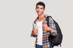 ser estudiante es un joven estudiante afroamericano genial y apuesto que sostiene libros y estira la mano con el pulgar hacia arriba mientras se enfrenta a un fondo gris foto