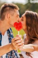 Stealing a kiss behind the lollipop.  Loving couple hiding behind red heart shaped lollipop while both standing outdoors photo