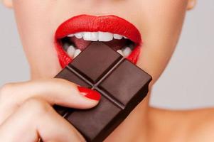 Indulging in a sweet affair with chocolate. Close-up of a beautiful women holding finger on her red lips while standing against grey background photo