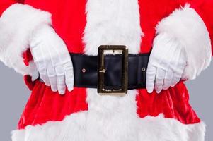 santa está aquí cerca del tradicional santa claus ajustando su cinturón mientras está de pie contra un fondo gris foto