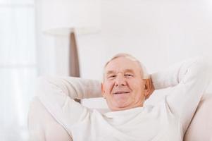 Relaxing at home. Happy senior man holding hands behind head and smiling while sitting in chair photo