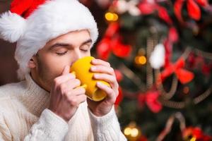 Hot drink at winter night. Handsome young man in Santa hat drinking hot drink with Christmas Tree in the background photo