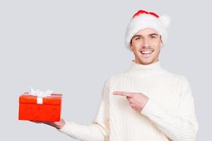 The best Christmas present. Cheerful young man in Santa hat holding a gift box and pointing it while standing against grey background photo