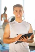 Confident instructor. Handsome young man holding clipboard and smiling while woman running on treadmill in the background photo