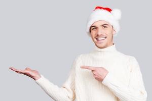 Your advertising here. Cheerful young man in Santa hat holding copy space and pointing it while standing against grey background photo