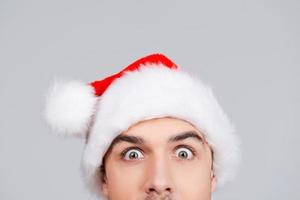 No way Surprised young man in Santa hat staring at you while standing against grey background photo