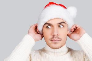 Feeling suspicious about Surprised young man looking away and adjusting his Santa hat while standing against grey background photo