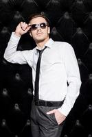 Making business look good. Handsome young man in shirt and tie adjusting his sunglasses and looking away while standing against black background photo