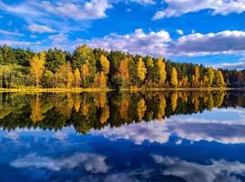 Autumn colored forest and its reflection in water with a blue cloudy sky. yellow and orange trees, green pine. Scenery photo