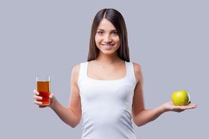 Apple or apple juice Beautiful young woman holding an apple in one hand and glass with juice in another while standing against grey background photo