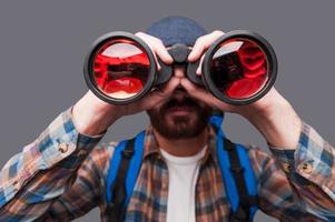 Discovering new places. Confident young bearded man carrying backpack and looking through binoculars while standing against grey background photo