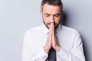 Praying for success. Portrait of concentrated mature man in shirt and tie holding hands clasped near face and keeping eyes closed while standing against grey background photo