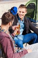 Cheers to friendship Top view of two handsome young men cheering with beer and smiling while sitting near the tent and on the sand photo