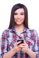 Typing message to friend. Beautiful young woman holding mobile phone and smiling at camera while standing isolated on white photo