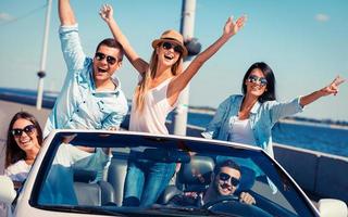 Friends in convertible. Group of young happy people enjoying road trip in their white convertible and raising their arms photo