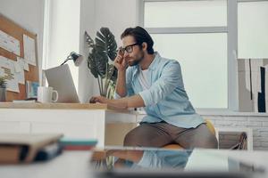 Confident young man using laptop while sitting at his working place in the office photo