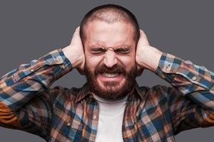 Too loud sound. Furious young bearded man covering ears with hands and keeping eyes closed while standing against grey background photo