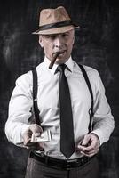 Money is not a problem. Serious senior man in hat and suspenders smoking cigar and stretching out money while standing against dark background photo