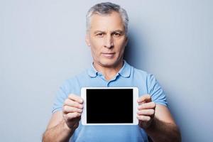Copy space on his tablet. Confident senior man showing his digital tablet and smiling while standing against grey background photo