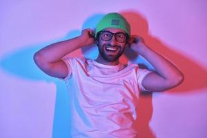 joven feliz ajustando su sombrero y sonriendo mientras está de pie contra un fondo colorido foto