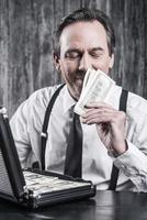 I love a smell of money. Satisfied senior man in shirt and suspenders sitting at the table and smelling money while case full of paper currency laying near him photo