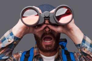 Surprised young bearded man carrying backpack and looking through binoculars while standing against grey background photo