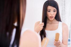 Hair loss. Depressed young woman looking at her hairbrush and expressing negativity while standing against mirror photo