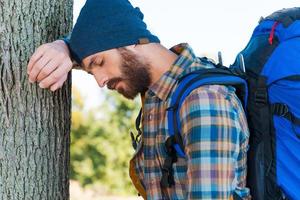 Tired traveler. Side view of tired young man with backpack leaning at the tree and keeping eyes closed photo