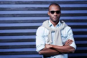 Elegance and style. Handsome young African man in sunglasses keeping arms crossed and looking at camera while standing against striped background photo