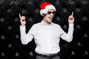 Are you ready to party Handsome young man in sunglasses and Santa hat pointing up and expressing positivity while standing against black background photo