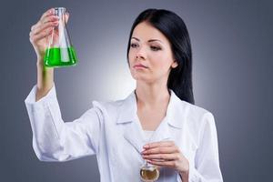 Research studies.  Beautiful  female doctor in white uniform holding flasks while standing against grey background photo