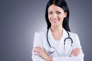 Your health is in the good hands. Confident female doctor in white uniform looking at camera and smiling while standing against grey background photo