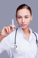 The benefit of medical advances. Confident female doctor in white uniform holding syringe and looking at it while standing against grey background photo