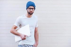 confiado y creativo. apuesto joven con anteojos sosteniendo una laptop y mirando la cámara mientras está de pie contra la pared de ladrillo foto