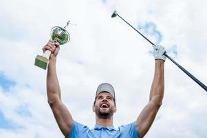 feliz ganador. vista en ángulo bajo de un joven golfista feliz sosteniendo un conductor y un trofeo mientras levanta los brazos con el cielo azul como fondo foto