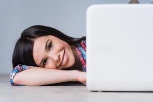 belleza de la era digital. mujer joven atractiva mirando fuera de la computadora portátil y sonriendo mientras está acostado en el piso de madera dura foto