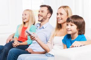 viendo su programa favorito. vista lateral de una familia feliz de cuatro personas que se unen y sonríen mientras ven la televisión juntos en casa foto