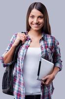 New age for studying. Happy young women carrying a backpack on her shoulder and holding digital tablet while standing against grey background photo