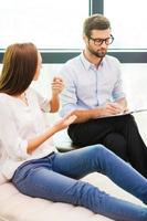 Sharing problems with professional. Worried young woman sitting at the chair and gesturing while male psychiatrist sitting close to her and writing something in his clipboard photo