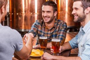 conocer viejos amigos. tres jóvenes felices sentados juntos en un pub de cerveza mientras dos de ellos se dan la mano foto