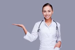 Copy space on her hand. Confident female doctor in white uniform holding copy space and smiling while standing against grey background photo