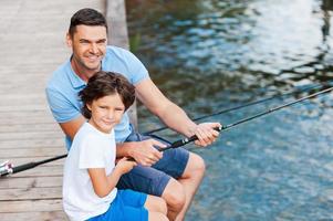 Spending great time together. Top view of father and son looking at camera and smiling while sitting at the quayside with fishing rods laying near them photo