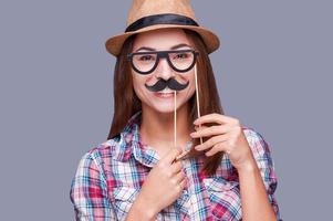 Cheerful fake face. Cheerful young woman in funky hat holding fake mustache and fake glasses on her face and looking at camera while standing against grey background photo